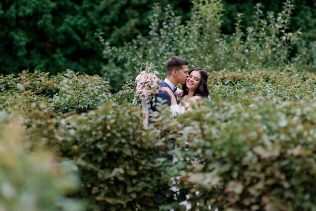 幸せな結婚式のカップルは笑顔と高い緑の茂みの中でキス