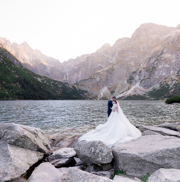 Счастливая свадьба пара стоит перед озером в окружении гор на огромной скале