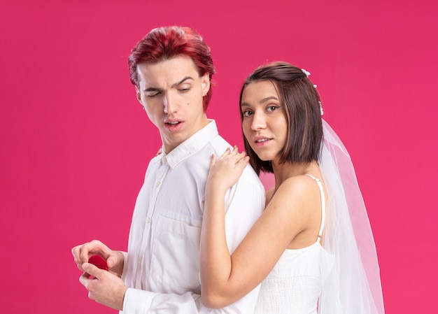 Happy wedding couple of posing together, man holding red box with wedding ring and his bride in white wedding dress standing behind over pink wall