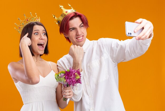 オレンジ色の壁の上に立っているスマートフォンを使用して元気に自分撮りをしている金の王冠を身に着けているウェディングドレスの花の花束と幸せな結婚式のカップル