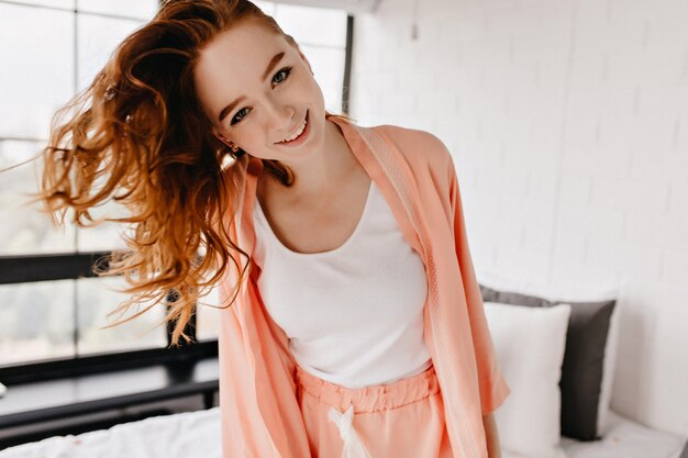 Счастливая белая девушка с улыбкой вьющихся волос. Вдохновленная рыжеволосая модель развлекается дома.