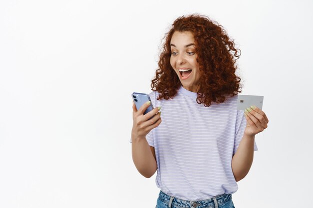 Happy winner looks at mobile phone. Excited redhead girl scream of joy, looking at smartphone screen, holding credit card, shopping online, white background.