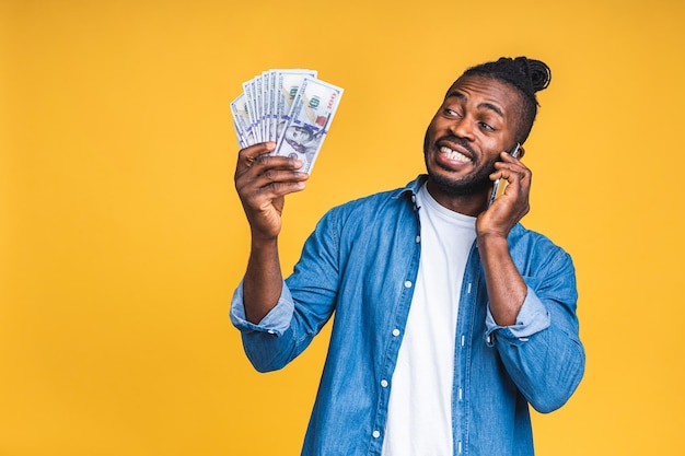Счастливый победитель! молодой богатый афро-американский темнокожий мужчина в непринужденной обстановке держит долларовые банкноты денег и мобильный телефон с сюрпризом, изолированным на желтом фоне. Premium Фотографии