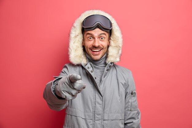 Happy winter time concept. Cheerful European man in outerwear indicates directly with glad expression sees something very pleasant in front.