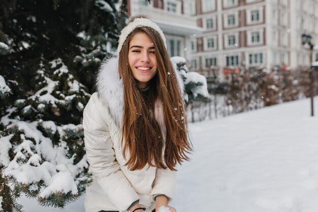 Happy winter time of positive pretty woman playing with snow. Smiling young woman with long brunette hair enjoying weekend during walk down street in frosty day..