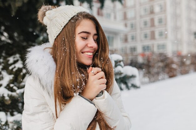 市で雪を楽しむ若いうれしそうな女性の幸せな冬時間。魅力的な女性、長いブルネットの髪、目を閉じて笑っています。