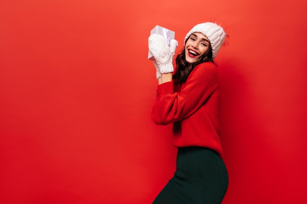 Happy woman in bright sweater and warm mittens holds white gift box excited girl with red lips poses on isolated background
