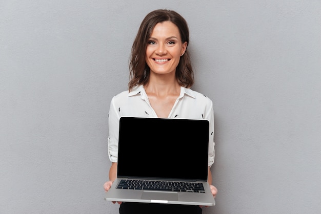 Счастливая женщина в деловой одежде, показывая пустой экран ноутбука на сером