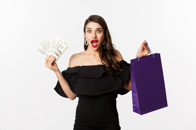 Happy woman buyer holding shopping bag and money, standing in black dress over white background. Copy space