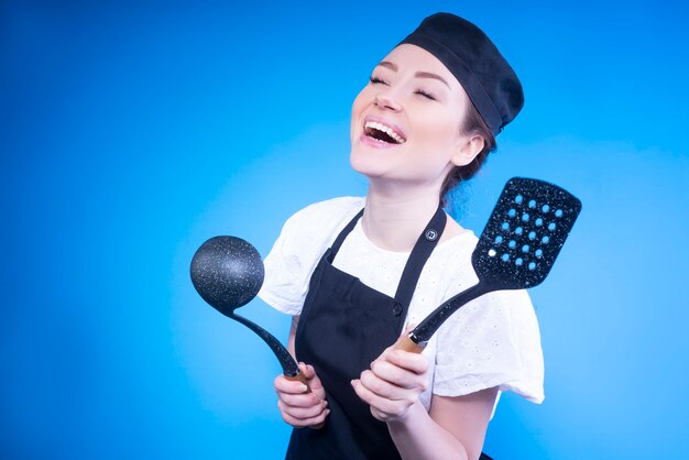 Счастливый шеф-повар женщина смеется и держит кухонную утварь в руках против синей стены