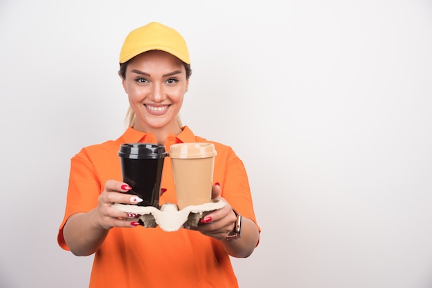 Happy woman courier holding two cups of coffees on white wall. 