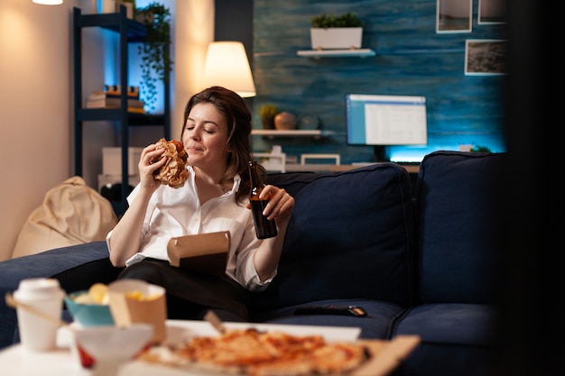 Happy woman eating tasty delicious delivery burger relaxing on sofa watching comedy movie