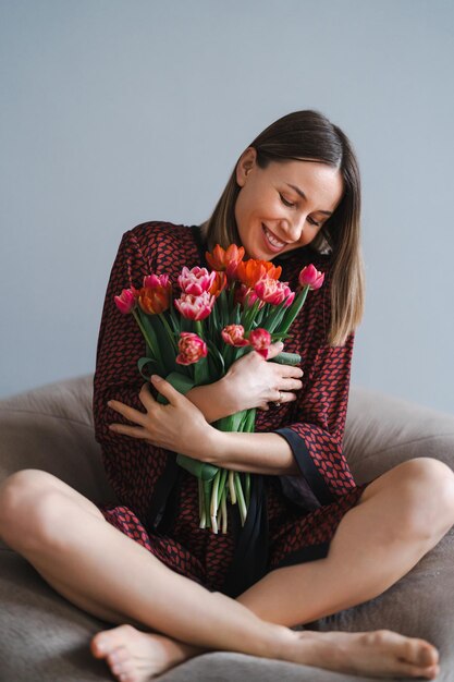 幸せな女性はチューリップの花束を楽しむ快適なビーンバッグでリラックスしながら花の束を楽しむ主婦甘い家アレルギー無料