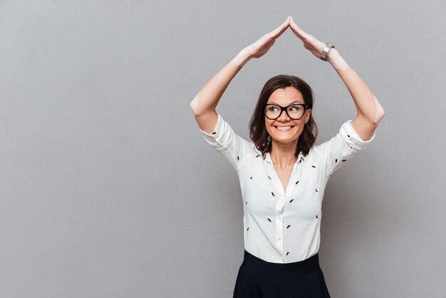 Happy woman in eyeglasses and business clothes hiding under hands like home on gray