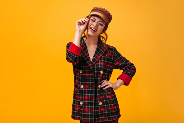 Happy woman in eyeglasses, cap and plaid jacket smiling on orange wall