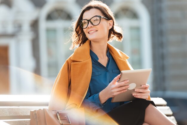Happy woman in eyeglasses holding tablet computer and looking away