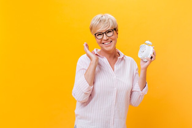 Happy woman in eyeglasses and pink outfit poses with alarm clock on orange background