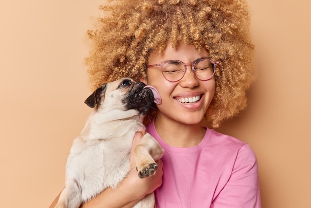 Happy woman gets kiss from favorite pet smiles gladully keeps eyes closed from pleasure Pug dog licks owner expresses love People domestic animals responsibility and relatioonship concept