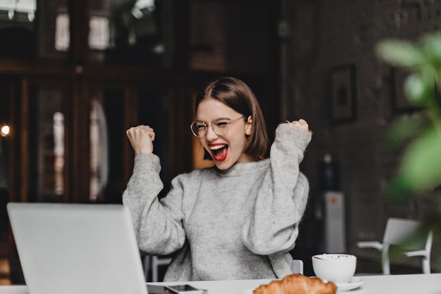 Happy woman in glasses makes winning gesture and sincerely rejoices. Lady with red lipstick dressed in gray sweater looking at laptop.