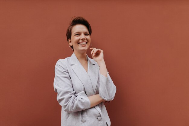 Happy woman in grey jacket laughing on brown background. Cheerful young girl in oversize stylish suit smiles and poses on isolated