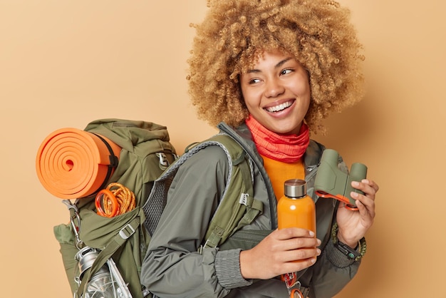 Happy woman hiker enjoys camping time carries big rucksack holds bottle with fresh water and binoculars going to climb mountain explores new places leads active lifestyle looks gladfully away