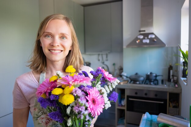 Happy woman holding bunch of flowers, posing in home kitchen, looking at camera and smiling. Women day or special date concept