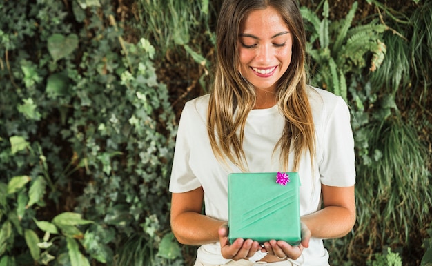 Foto gratuita donna felice che tiene il contenitore di regalo verde davanti al fondo delle foglie verdi