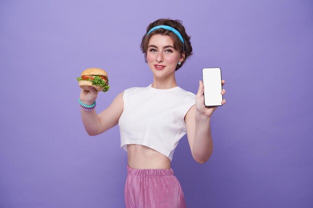 Happy woman holding hamburger and showing food delivery app on mobile phone to order lunch