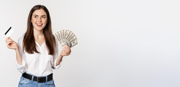 Free photo happy woman holding money and credit card with thoughtful face concept of loan and microcredit stand