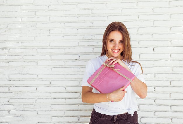 Happy woman holding pink gift box in front of brick wall