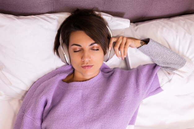 Donna felice a casa sul letto comodo indossando pullover vestiti caldi, ascoltare musica