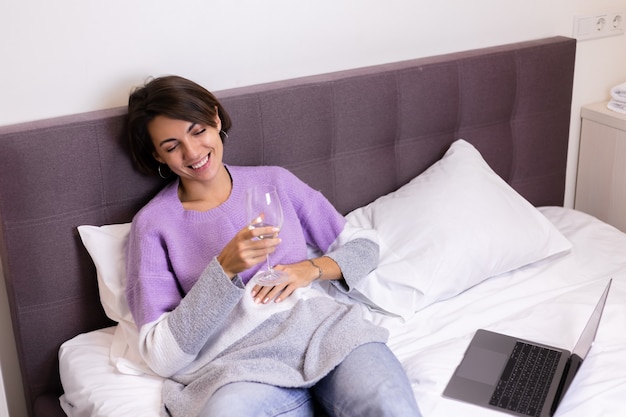 Счастливая женщина в теплом пуловере в постели с бокалом вина одна отдыхает, смотря комедию, смех фильма