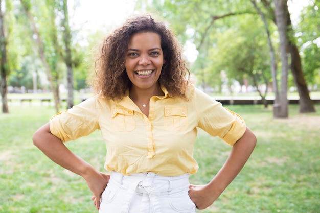 Happy woman keeping hands on hips and posing at camera in park