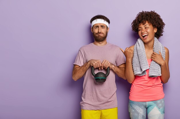 Happy woman laughs sincerely, points away with thumb, indicates at boyfriend who lifts weight