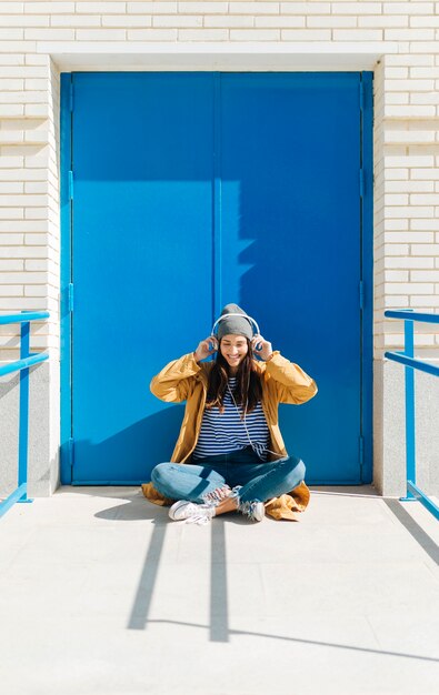 happy woman listening music on smart phone sitting  against blue door outdoors