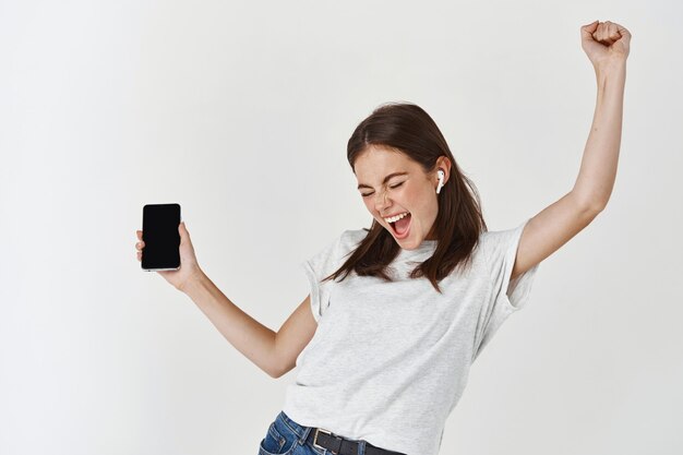 Happy woman listening to music in wireless headphones and holding smartphone, dancing and singing over white wall