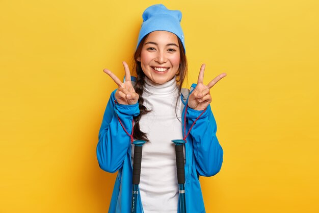 Счастливая женщина делает жест мира, позирует с треккинговыми палками, одетая в синюю шляпу и куртку, любит пешие прогулки, с удовольствием смотрит в камеру, изолирована над желтой стеной
