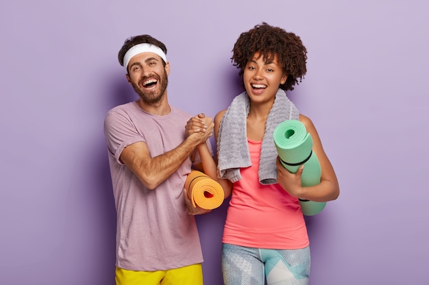 Happy woman and man keep hands together, dressed in sportwear, hold fitness mats