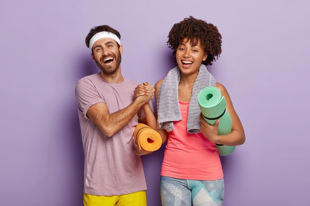 Happy woman and man keep hands together, dressed in sportwear, hold fitness mats