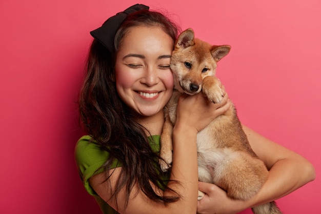 행복한 여자 애완 동물 소유자는 얼굴 근처에 귀여운 강아지를 보유하고, 시바 inu 강아지를 운반하고, 즐겁게 미소를 짓고, 분홍색 배경 위에 고립 된 달콤한 순간을 즐깁니다.