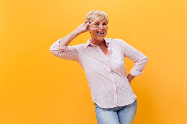 Happy woman in pink outfit smiles and shows peace sign on orange background