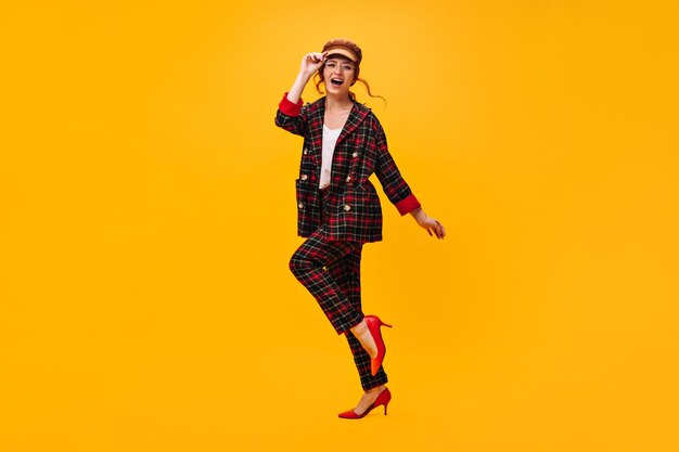 Happy woman in plaid outfit moves on orange background Cheerful girl in brown cap and eyeglasses has fan on isolated backdrop