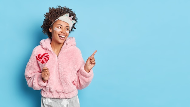 Happy woman points away on copy space feels energized in morning after good sleep indicates aside and advertises product for great nap holds lollipop wears pajama isolated over blue wall