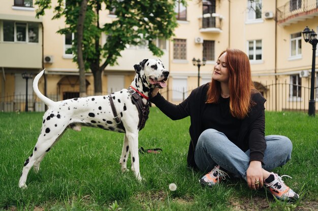 都会の街歩き中に緑の芝生に座ってダルメシアン犬とポーズをとって遊んで幸せな女性