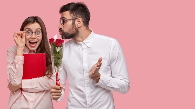 Happy woman recieves red roses from handsome guy, looks positively through spectacles