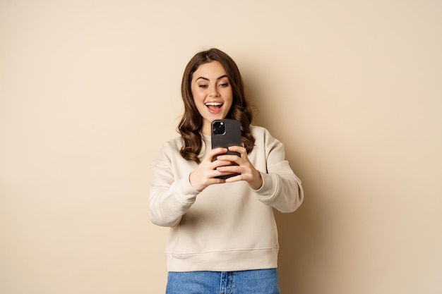 Happy woman recording video shooting photo on smartphone camera and smiling standing over beige back...