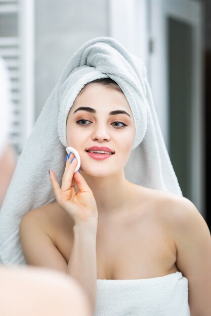 Счастливая женщина, снимающая макияж, очищающие салфетки, лосьон, зеркало в ванной комнате