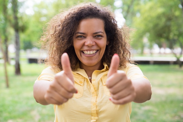 Happy woman showing thumbs up and posing at camera in park