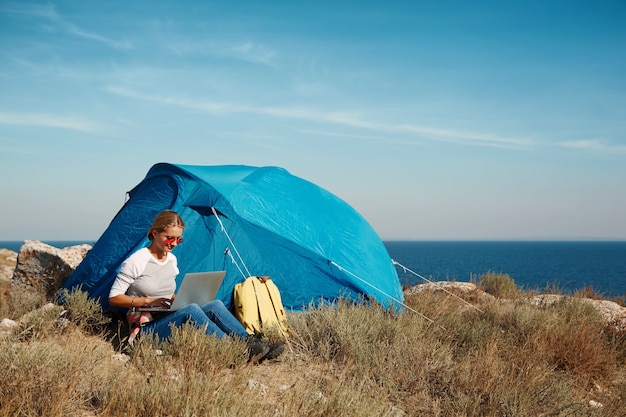 Счастливая женщина, сидящая возле палатки с ноутбуком