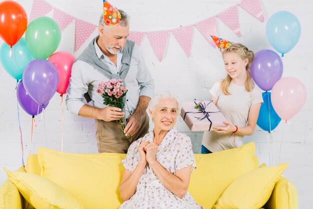 Happy woman sitting on sofa in front of husband and granddaughter holding birthday gifts